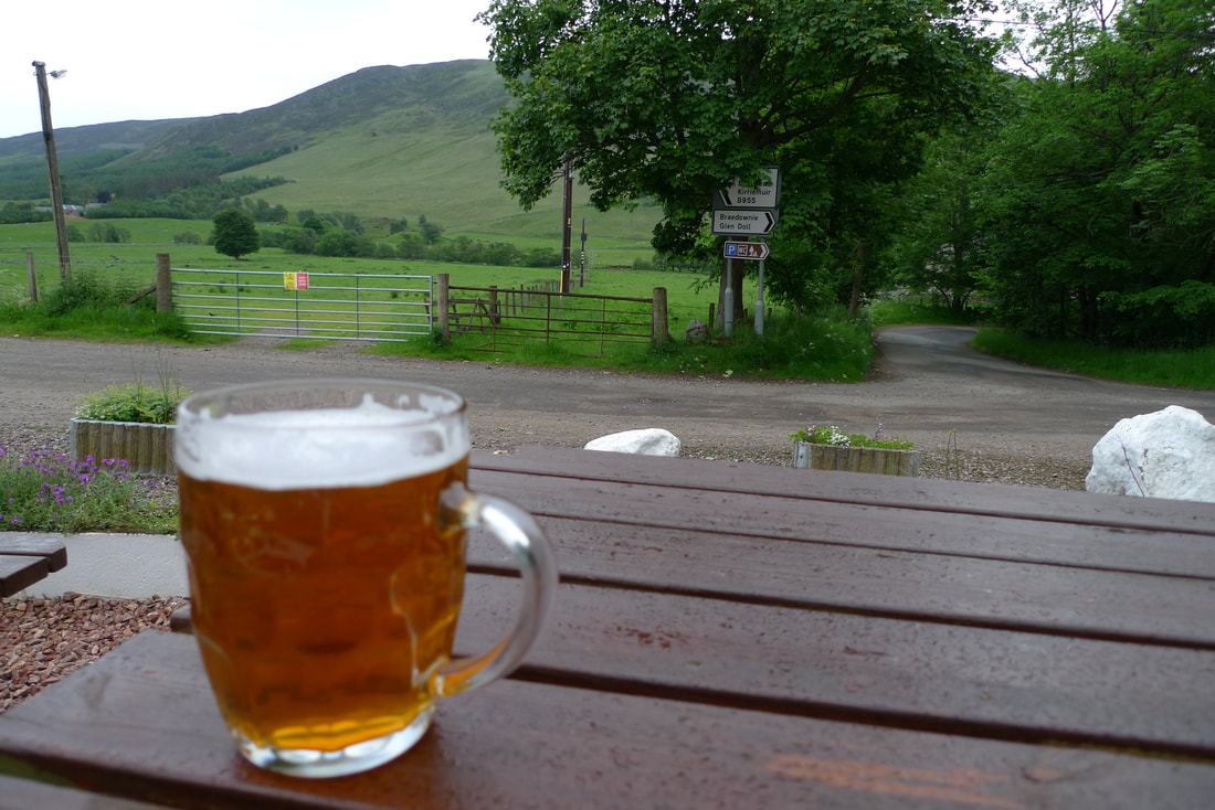 A pint of Glen Clova Ale on a table outside the Glen Clova Hotel. There is a view of hills.of the Glen Clova Hotel